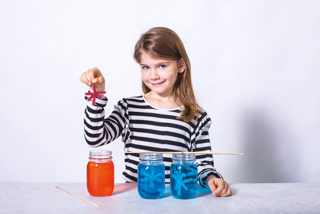 Fille qui tient un flocon de sucre devant des bocaux avec une solution bleu et une solution orange.